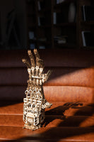 Een WoodTrick Cyber Hand - Robot Hand Bouwpakket van WoodTrick staat rechtop op een bruine leren bank, zonlicht accentueert de gedetailleerde tandwielen en gewrichten. De vage boekenplanken op de achtergrond geven het geheel een gezellig tintje.