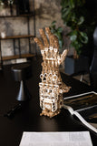 Het WoodTrick Cyber Hand - Robot Hand Bouwpakket van WoodTrick, met beweegbare vingers, wordt tentoongesteld op een zwarte tafel met documenten, een open tijdschrift, een zonnebril en een kruk in de buurt, tegen een achtergrond van planten.