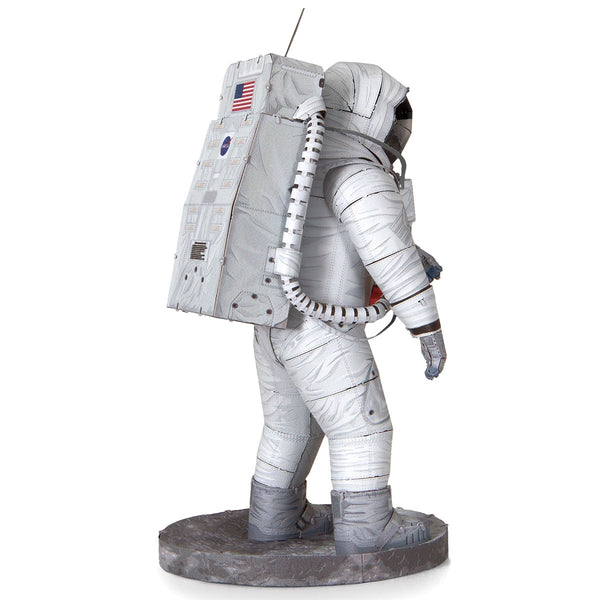 De SlimSpul Apollo 11 astronaut - metalen bouwpakket is een levensgroot model met een astronaut in een wit pak met een Amerikaanse vlag patch, gouden vizier helm, grote rugzak, en staande op een ronde grijze platform, gezien vanaf de zijkant.