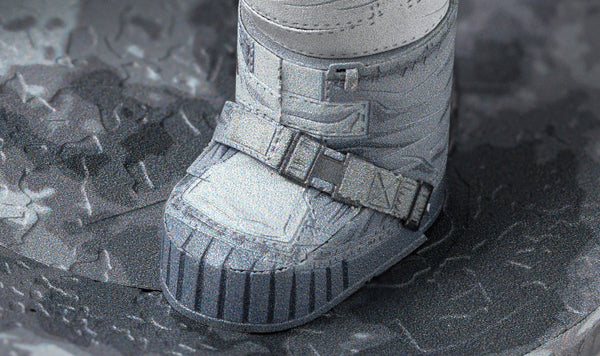 Een close-up van de laars van een Apollo 11-astronaut op het stoffige maanoppervlak is de inspiratiebron voor het Apollo 11 astronaut - metalen bouwpakket van SlimSpul, compleet met gedetailleerde riemen en treden op grijs, rotsachtig terrein.