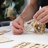 Een persoon zet de Sky Lodge DIY houten modelbouwkit van Tiny Treehouses Europe in elkaar, waarbij lasergesneden stukken in elkaar worden gezet op een witte tafel met houten platen en kleine LED-lampjes verspreid in de buurt.