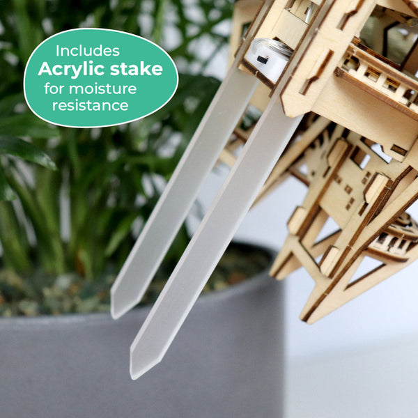 Close-up van de Horizons Edge bloempot van Tiny Treehouses Europe met een houten structuur en twee witte acrylstaken in de grond. Op een groen label staat: "Inclusief acrylstokken voor vochtbestendigheid. Plantenbladeren verschijnen op de achtergrond.
