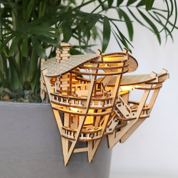 Horizons Edge van Tiny Treehouses Europe is een klein, ingewikkeld luxe boomhutmodel dat vastzit aan een grijze bloempot. Het is gemaakt van een zelfgemaakt houten bouwpakket en straalt van binnenuit, tegen groene plantenbladeren op de achtergrond.