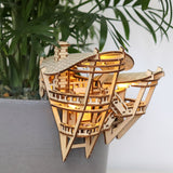 Horizons Edge van Tiny Treehouses Europe is een klein, ingewikkeld luxe boomhutmodel dat vastzit aan een grijze bloempot. Het is gemaakt van een zelfgemaakt houten bouwpakket en straalt van binnenuit, tegen groene plantenbladeren op de achtergrond.