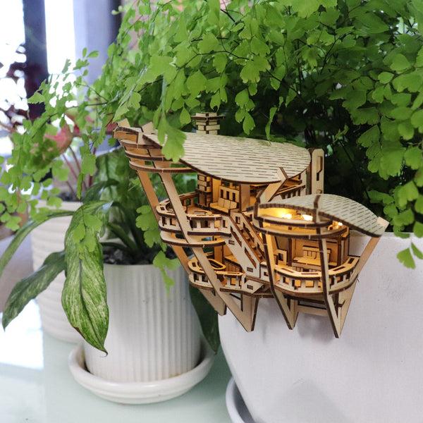Horizons Edge van Tiny Treehouses Europe - een klein, ingewikkeld doe-het-zelf houten model van een gebouw - wordt vastgeklikt aan de rand van een witte plantenbak gevuld met weelderige groene kamerplanten en straalt van binnenuit.