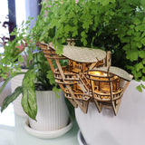 Horizons Edge van Tiny Treehouses Europe - een klein, ingewikkeld doe-het-zelf houten model van een gebouw - wordt vastgeklikt aan de rand van een witte plantenbak gevuld met weelderige groene kamerplanten en straalt van binnenuit.