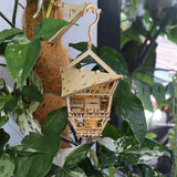 De Sky Lodge van Tiny Treehouses Europe is een klein, gedetailleerd doe-het-zelf houten huisje met zichtbare kamers en ingewikkelde architectuur, hangend tussen weelderige planten aan een houten arm op een met kokosvezel omwikkelde paal.