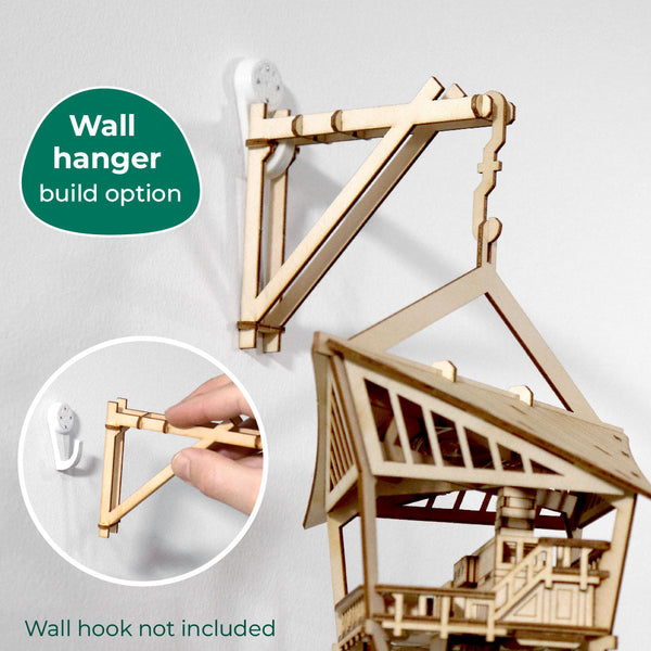 Een houten muurhanger ondersteunt het Sky Lodge DIY model van Tiny Treehouses Europe, gemonteerd op een witte muur; een hand bevestigt de hanger aan een muurhaak. Tekst: Muurhanger bouwoptie. Muurhaak niet inbegrepen.