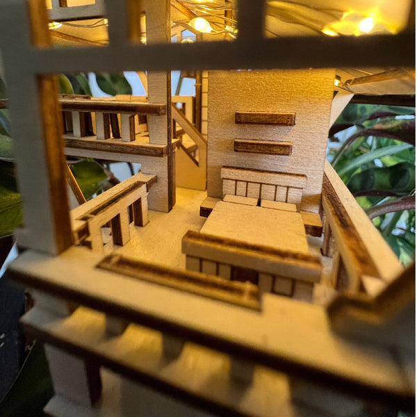 Close-up van Tiny Treehouses Europe's Tropical Lookout DIY houten modelbouwpakket, met tafels en banken verlicht door warme LED-verlichting. Groene bladeren op de achtergrond creëren een gezellige sfeer in de jungle.