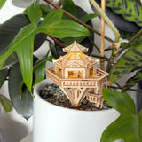 De Woodland Outpost van Tiny Treehouses Europe is een gedetailleerd houten modelbouwpakket dat op kiezels in een witte plantenbak ligt, omringd door groene planten - een charmant doe-het-zelf tafereel voor thuis.