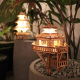 Woodland Outpost van Tiny Treehouses Europe is een doe-het-zelf houten modelbouwpakket met een miniatuur boomhut met oplichtende LED-lampjes, genesteld in een plantenpot met witte stenen en groen gebladerte voor een warme, uitnodigende verlichting.