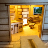 Een close-up van het Woodland Outpost DIY houten modelbouwpakket van Tiny Treehouses Europe, met miniatuurtafels en kasten die warm worden verlicht door LED's aan de binnenkant en bekeken worden door een open deuropening.