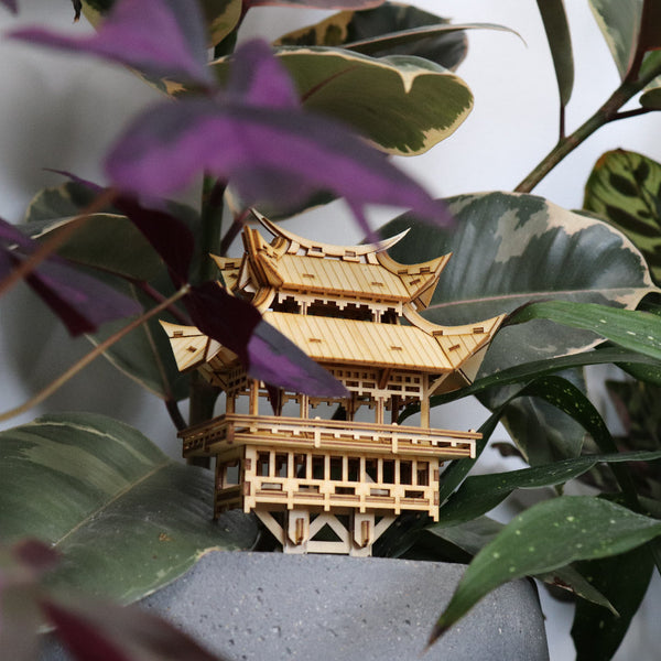De Temple of Gratitude DIY houten model van Tiny Treehouses Europe staat op een grijze plantenbak, omringd door groene en paarse kamerplantenbladeren.
