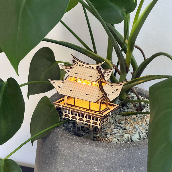 De Tempel van Dankbaarheid van Tiny Treehouses Europe is een klein, verlicht doe-het-zelf houten model van een traditioneel Japans gebouw, genesteld tussen groene bladeren en kiezels in een grijze pot en versterkt met LED-verlichting.