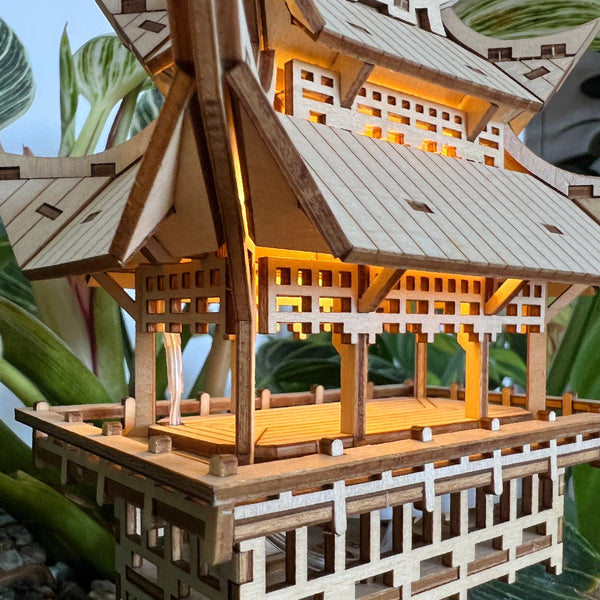 Een close-up van de Temple of Gratitude van Tiny Treehouses Europe, een gedetailleerd houten bouwpakket dat lijkt op een pagode in Japanse stijl, warm verlicht door LED's met groene bladplanten op de achtergrond.