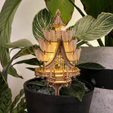 De Temple of Joy van Tiny Treehouses Europe is een kleine, gedetailleerde DIY pagode van houten modelbouwpakket met verlichte ramen, uitgestald in een zwarte bloempot en geplaatst tegen groene bladplanten.