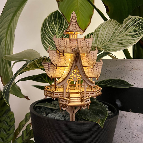 De Temple of Joy van Tiny Treehouses Europe is een kleine, gedetailleerde DIY pagode van houten modelbouwpakket met verlichte ramen, uitgestald in een zwarte bloempot en geplaatst tegen groene bladplanten.