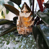De Temple of Joy van Tiny Treehouses Europe is een klein, ingewikkeld houten pagode-model met zachte LED-verlichting, genesteld tussen weelderige groene plantenbladeren en wijnranken.