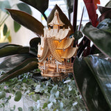 De Temple of Joy van Tiny Treehouses Europe is een klein, ingewikkeld houten pagode-model met zachte LED-verlichting, genesteld tussen weelderige groene plantenbladeren en wijnranken.