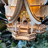 Een close-up van de Temple of Joy DIY houten modelbouwkit van Tiny Treehouses Europe, met ingewikkeld houtsnijwerk en leuningen tussen groene bladeren. Warme LED-lampjes schijnen vanuit de binnenkant van het bouwwerk.