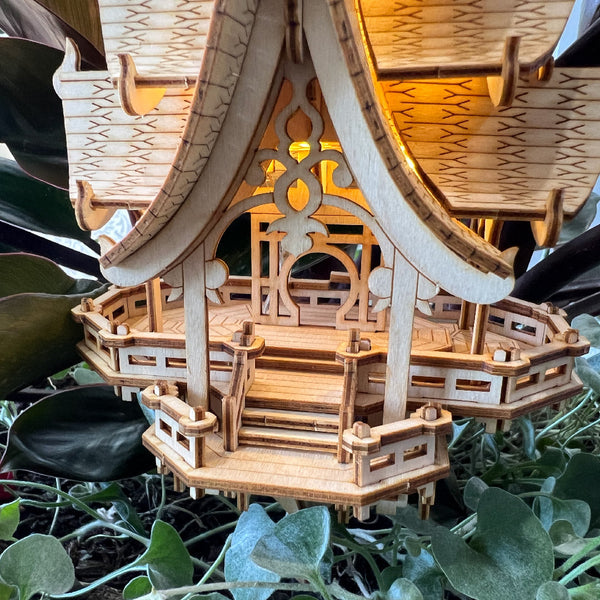 Een close-up van de Temple of Joy DIY houten modelbouwkit van Tiny Treehouses Europe, met ingewikkeld houtsnijwerk en leuningen tussen groene bladeren. Warme LED-lampjes schijnen vanuit de binnenkant van het bouwwerk.