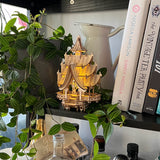 De Tempel van Vreugde, een klein verlicht houten model van Tiny Treehouses Europe, staat op een plank naast bladerranken en kookboeken als Simply Nigella en The Vintage Tea Party Book, met LED-lampjes die een gezellige gloed verspreiden.