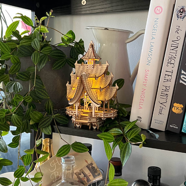 De Tempel van Vreugde, een klein verlicht houten model van Tiny Treehouses Europe, staat op een plank naast bladerranken en kookboeken als Simply Nigella en The Vintage Tea Party Book, met LED-lampjes die een gezellige gloed verspreiden.