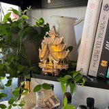 De Tempel van Vreugde, een klein verlicht houten model van Tiny Treehouses Europe, staat op een plank naast bladerranken en kookboeken als Simply Nigella en The Vintage Tea Party Book, met LED-lampjes die een gezellige gloed verspreiden.