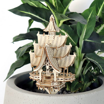 De Temple of Joy van Tiny Treehouses Europe, een gedetailleerd zelfgemaakt houten model pagode met gelaagde daken en decoratieve patronen, staat in een grijze plantenbak met groene bladplanten op de achtergrond.