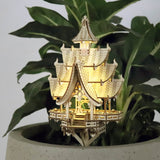 De Temple of Joy van Tiny Treehouses Europe, een gedetailleerd verlicht houten pagode-model uit een doe-het-zelf bouwpakket, staat op een grijze plantenbak te midden van groene bladeren en zachte LED-verlichting op de achtergrond.