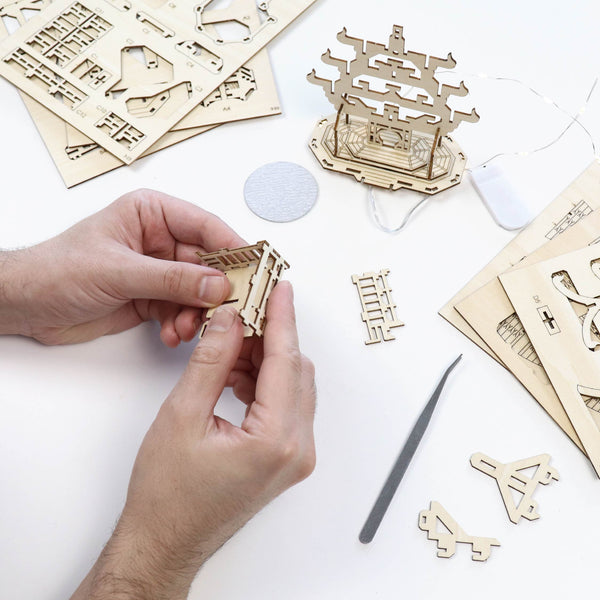 Een persoon zet de Temple of Joy DIY modelbouwkit van Tiny Treehouses Europe in elkaar op een wit oppervlak, met lasergesneden houten stukken, gereedschap, LED-verlichting en een gedeeltelijk gebouwde pagode zichtbaar.