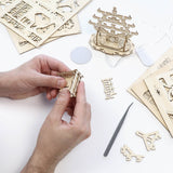Een persoon zet de Temple of Joy DIY modelbouwkit van Tiny Treehouses Europe in elkaar op een wit oppervlak, met lasergesneden houten stukken, gereedschap, LED-verlichting en een gedeeltelijk gebouwde pagode zichtbaar.