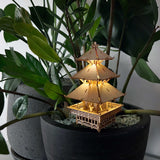 De Tempel van Sereniteit van Tiny Treehouses Europe - een klein, verlicht houten pagode-model - staat tussen bladerrijke planten in een zwarte pot met witte kiezels, met zachte LED-lampjes die oplichten, naast een andere, soortgelijke pot tegen een effen achtergrond.