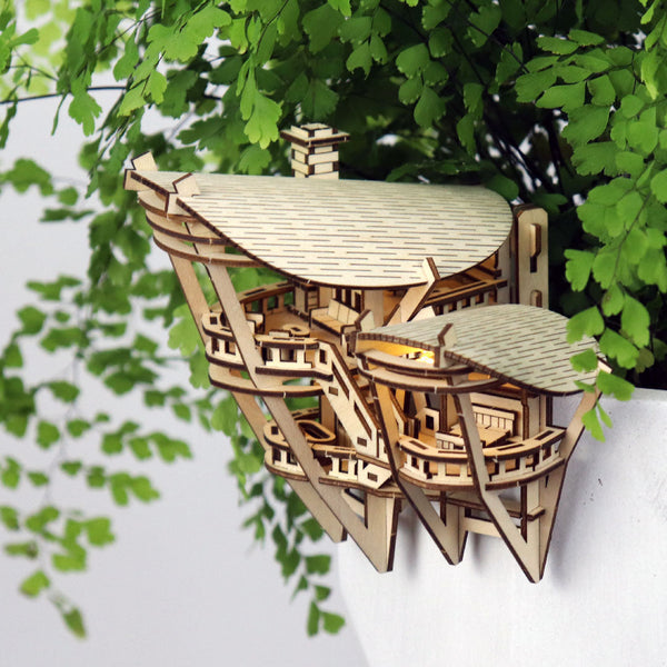 Horizons Edge van Tiny Treehouses Europe is een klein, ingewikkeld gemaakt doe-het-zelf houten model van een modern gebouw, vastgeklikt aan de rand van een witte plantenbak met groene bladplanten op de achtergrond.