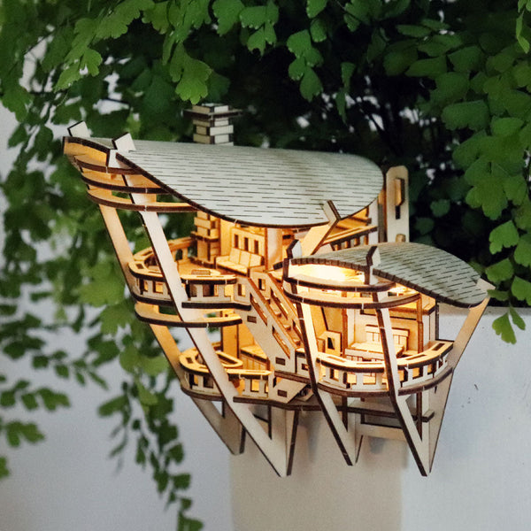 Horizons Edge van Tiny Treehouses Europe is een klein, gedetailleerd model van een luxe boomhut met binnenverlichting tegen bladerrijke planten en een witte muur - een ideale keuze voor liefhebbers van doe-het-zelf houten modelbouwpakketten.