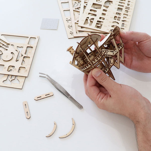 Handen die het Horizons Edge DIY houten modelbouwpakket van Tiny Treehouses Europe in elkaar zetten, met lasergesneden houten platen, kleine onderdelen en een pincet op een witte tafel.