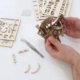 Handen die het Horizons Edge DIY houten modelbouwpakket van Tiny Treehouses Europe in elkaar zetten, met lasergesneden houten platen, kleine onderdelen en een pincet op een witte tafel.