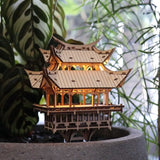 De Tempel van Dankbaarheid van Tiny Treehouses Europe is een kleine, ingewikkeld gedetailleerde DIY houten pagode model kit met warme LED-verlichting, uitgestald in een grijze plantenbak omringd door weelderige groene planten.