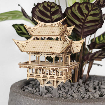 De Tempel van Dankbaarheid van Tiny Treehouses Europe, een klein doe-het-zelf bouwpakket van hout, wordt tentoongesteld in een grijze pot met kiezels tegen groene en paarse planten.