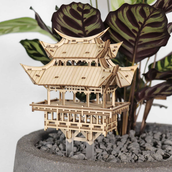 De Tempel van Dankbaarheid van Tiny Treehouses Europe, een klein doe-het-zelf bouwpakket van hout, wordt tentoongesteld in een grijze pot met kiezels tegen groene en paarse planten.