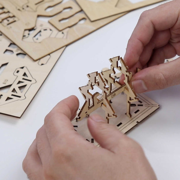 Iemand monteert kleine, in elkaar grijpende stukken van de Tempel van Dankbaarheid DIY houten modelbouwpakket van Tiny Treehouses Europe op een vlakke ondergrond, met meer ongemonteerde stukken van het pakket zichtbaar op de achtergrond.
