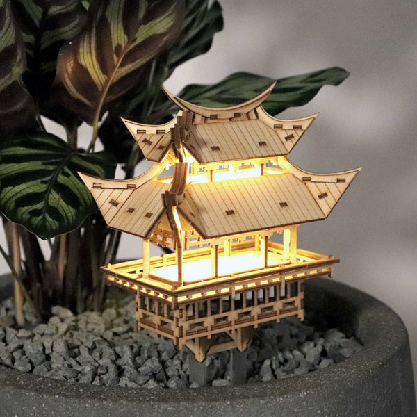 De Tempel van Dankbaarheid van Tiny Treehouses Europe is een klein, verlicht houten model van een pagode met meerdere verdiepingen, gebouwd van een doe-het-zelf bouwpakket, tentoongesteld in een pot met grijze kiezels en bladgroene planten op de achtergrond.