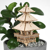 De Tempel van Sereniteit van Tiny Treehouses Europe, een klein doe-het-zelf houten modelbouwpakket van een pagode met drie verdiepingen, staat in een potplant met brede groene bladeren en grijze kiezels tegen een effen witte achtergrond.
