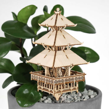 De Tempel van Sereniteit van Tiny Treehouses Europe, een klein doe-het-zelf houten modelbouwpakket van een pagode met drie verdiepingen, staat in een potplant met brede groene bladeren en grijze kiezels tegen een effen witte achtergrond.