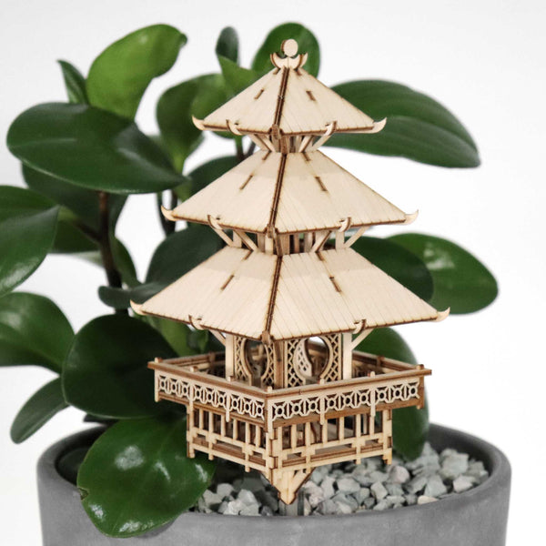 De Tempel van Sereniteit van Tiny Treehouses Europe, een klein doe-het-zelf houten modelbouwpakket van een pagode met drie verdiepingen, staat in een potplant met brede groene bladeren en grijze kiezels tegen een effen witte achtergrond.