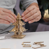 Een persoon zet de Tiny Treehouses Europe "Temple of Serenity" DIY houten modelbouwpakket in elkaar op een witte tafel, met extra houten onderdelen, LED-verlichting en montagegereedschap in de buurt.