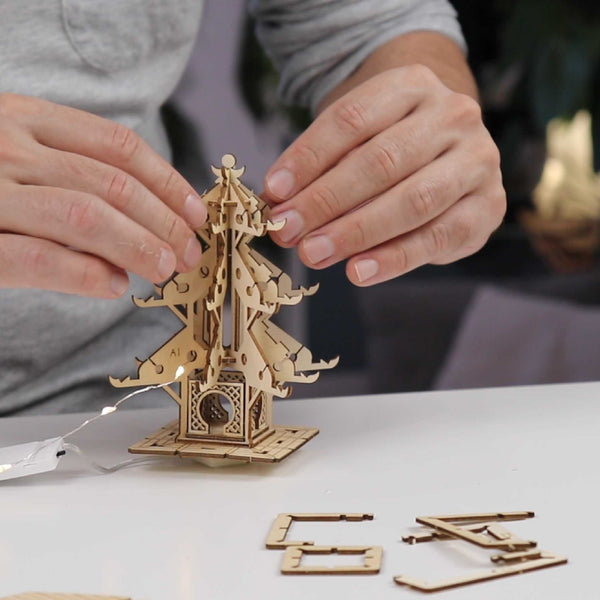 Een persoon zet de Tiny Treehouses Europe "Temple of Serenity" DIY houten modelbouwpakket in elkaar op een witte tafel, met extra houten onderdelen, LED-verlichting en montagegereedschap in de buurt.