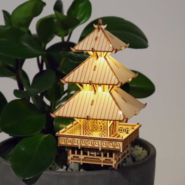De Tempel van Sereniteit van Tiny Treehouses Europe, een klein verlicht houten pagode-model uit een bouwpakket, rust op witte kiezels in een grijze plantenbak omringd door bladeren van kamerplanten.