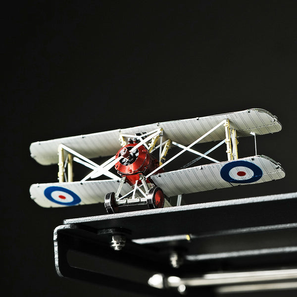Een close-up van het SlimSpul nederland b.v. Sopwith Camel jachtvliegtuig metalen bouwpakket, met een rode neus en witte vleugels met blauwe en rode rondellen, afgebeeld op een zwart vlak tegen een donkere achtergrond.