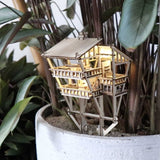 De Tropical Lookout van Tiny Treehouses Europe is een klein, verlicht DIY houten boomhutmodel geplaatst tussen groene bladeren in een grote witte plantenbak, met oplichtende LED-lampjes die een gezellig junglehuiseffect creëren in miniatuur gebladerte.